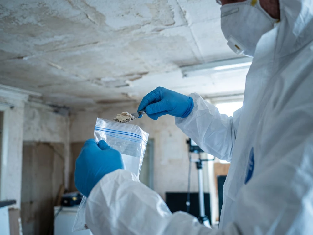 Surveyor in protective coveralls taking a material sample from a ceiling for asbestos analysis